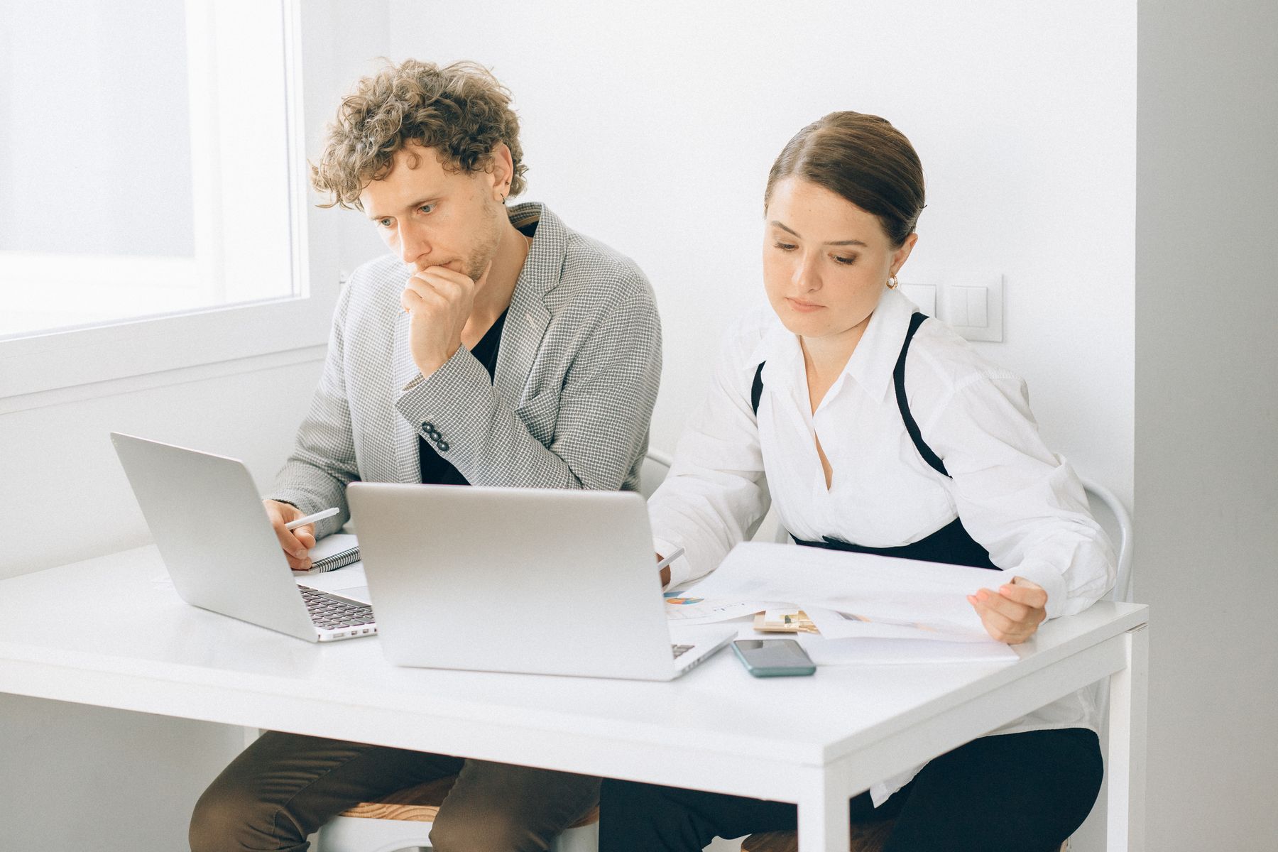 Two professionals reviewing laptops and printed plans at a bright desk.
